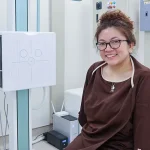 Dra. Bianca Battung wearing a medical gown sits in a diagnostic room at Shinagawa, ready for an X-ray or imaging procedure.
