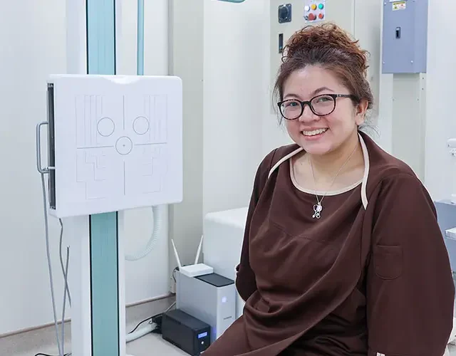 Dra. Bianca Battung wearing a medical gown sits in a diagnostic room at Shinagawa, ready for an X-ray or imaging procedure.
