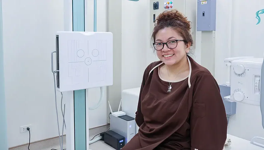 Dra. Bianca Battung wearing a medical gown sits in a diagnostic room at Shinagawa, ready for an X-ray or imaging procedure.