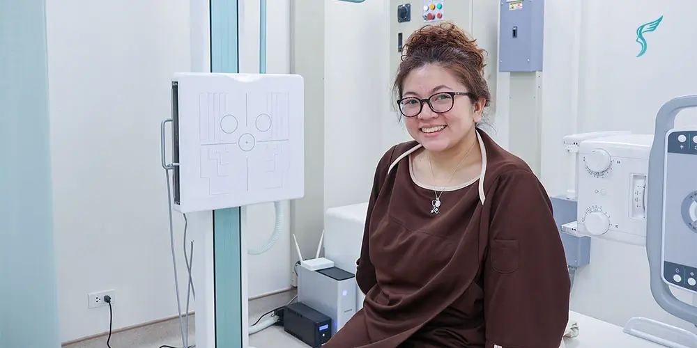 Dra. Bianca Battung wearing a medical gown sits in a diagnostic room at Shinagawa, ready for an X-ray or imaging procedure.