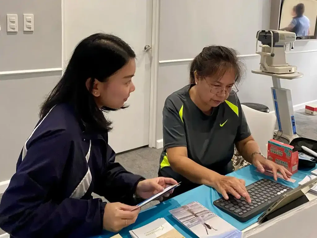 Staff assisting a patient with registration at a healthcare facility front desk, with brochures and medical equipment visible in the background.