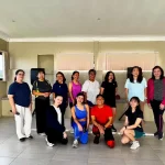 Group photo of fitness class participants in activewear smiling after a workout session inside a well-lit indoor studio with beige walls and large windows.
