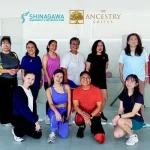 Group photo of fitness class participants in activewear smiling after a workout session inside a well-lit indoor studio with beige walls and large windows.
