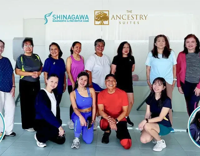 Group photo of fitness class participants in activewear smiling after a workout session inside a well-lit indoor studio with beige walls and large windows.