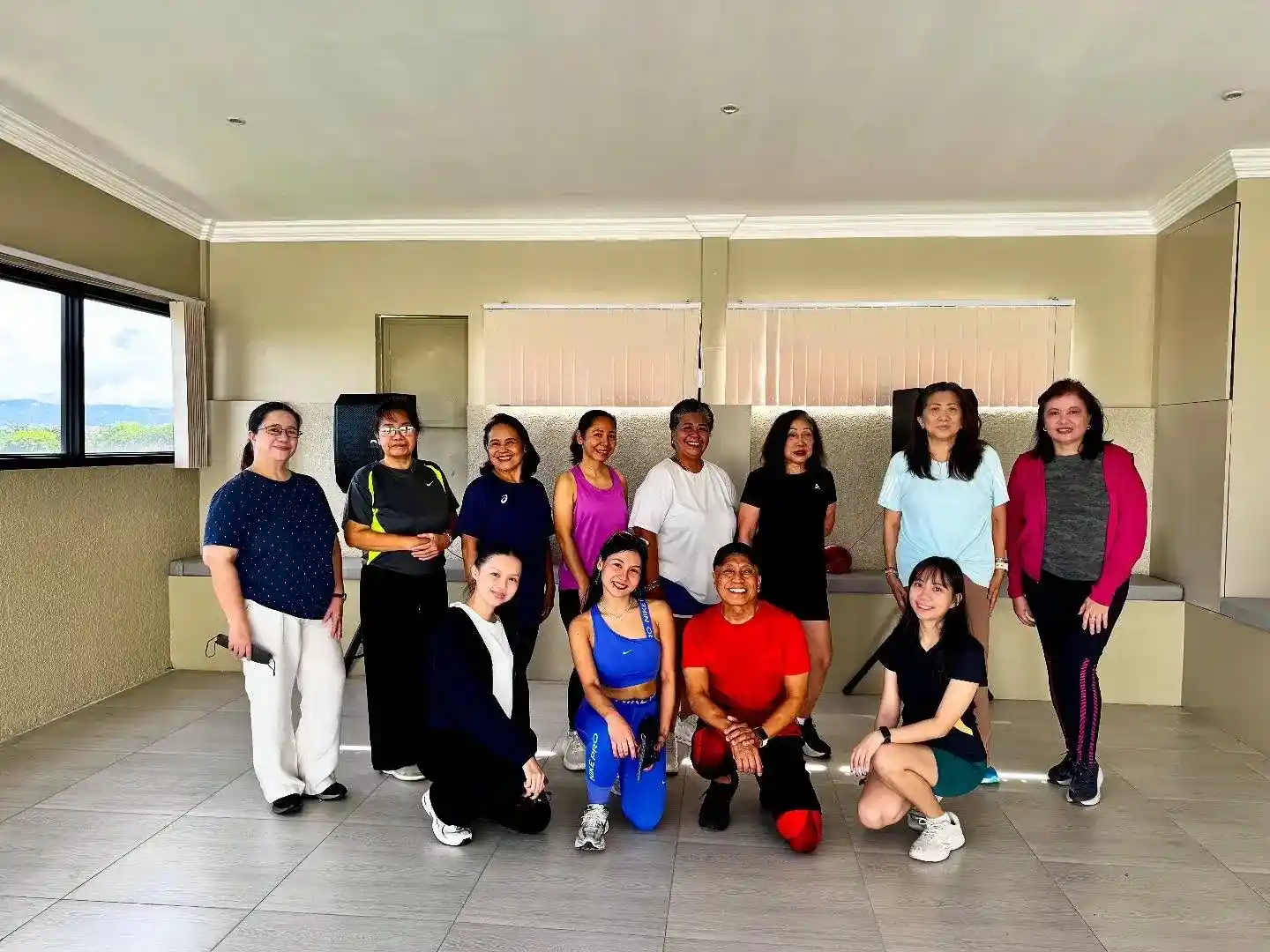 Group photo of fitness class participants in activewear smiling after a workout session inside a well-lit indoor studio with beige walls and large windows.