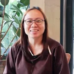 Female patient wearing glasses smiling during a video testimonial at Shinagawa Diagnostic & Preventive Care clinic, with indoor plants and natural lighting in the background.