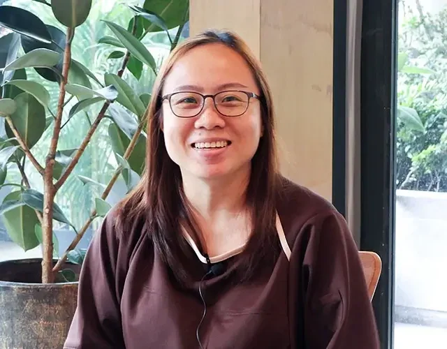 Female patient wearing glasses smiling during a video testimonial at Shinagawa Diagnostic & Preventive Care clinic, with indoor plants and natural lighting in the background.