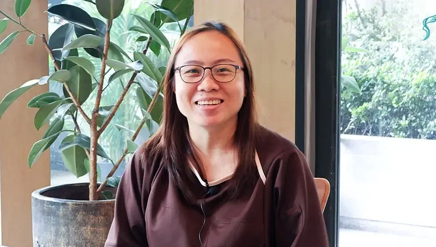 Female patient wearing glasses smiling during a video testimonial at Shinagawa Diagnostic & Preventive Care clinic, with indoor plants and natural lighting in the background.