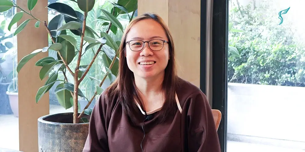 Female patient wearing glasses smiling during a video testimonial at Shinagawa Diagnostic & Preventive Care clinic, with indoor plants and natural lighting in the background.