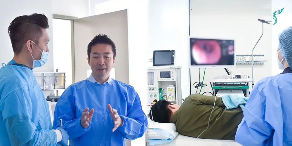 Medical team at Shinagawa Diagnostic & Preventive Care performing a colonoscopy procedure, with doctors in blue gowns and a patient lying on their side while the colon is displayed on a monitor.