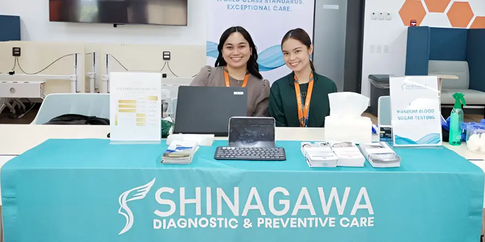 Shinagawa Diagnostic & Preventive Care representatives at an indoor health booth offering random blood sugar testing and wellness brochures at a corporate event.