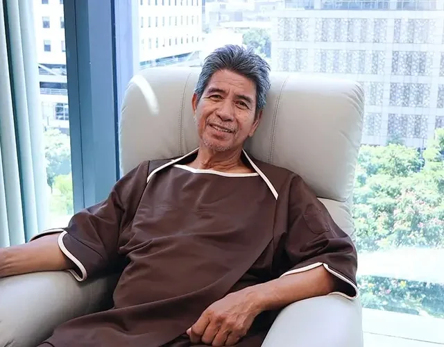 Elderly male patient in a medical gown relaxes in a recliner at Shinagawa Diagnostic & Preventive Care with city view behind.