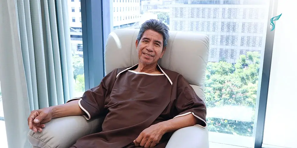 Elderly male patient in a medical gown relaxes in a recliner at Shinagawa Diagnostic & Preventive Care with city view behind.