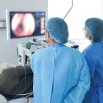 Medical team at Shinagawa Diagnostic & Preventive Care performing a colonoscopy procedure, with doctors in blue gowns and a patient lying on their side while the colon is displayed on a monitor.