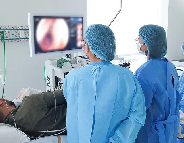 Medical team at Shinagawa Diagnostic & Preventive Care performing a colonoscopy procedure, with doctors in blue gowns and a patient lying on their side while the colon is displayed on a monitor.