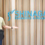 Male patient smiling and posing in front of the Shinagawa Diagnostic & Preventive Care clinic logo wall, showcasing a positive healthcare experience.
