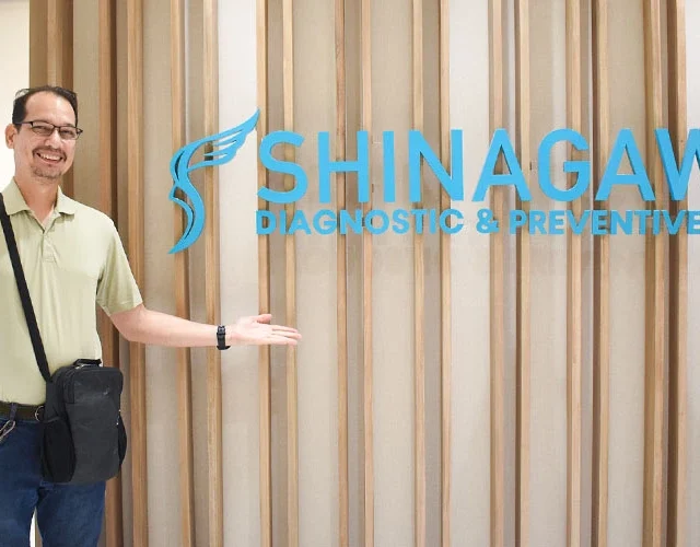 Male patient smiling and posing in front of the Shinagawa Diagnostic & Preventive Care clinic logo wall, showcasing a positive healthcare experience.