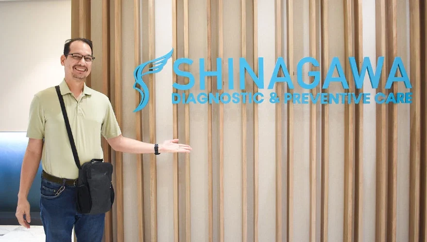 Male patient smiling and posing in front of the Shinagawa Diagnostic & Preventive Care clinic logo wall, showcasing a positive healthcare experience.